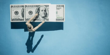 A wooden mannequin arm reaches toward a row of three hundred-dollar bills on a flat blue background, casting a shadow below. | 5StarsStocks