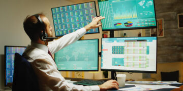 A man wearing a headset sits at a desk with multiple monitors displaying graphs, charts, and data. He is reaching up to touch one of the top screens, focusing on financial or business analytics. | 5StarsStocks