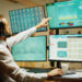 A man wearing a headset sits at a desk with multiple monitors displaying graphs, charts, and data. He is reaching up to touch one of the top screens, focusing on financial or business analytics. | 5StarsStocks