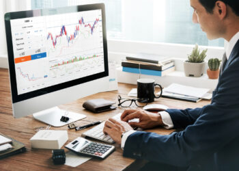 A person in a suit sits at a desk using a computer displaying financial charts and stock trading information, with a calculator, notebook, and coffee cup nearby. | 5StarsStocks