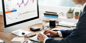 A person in a suit sits at a desk using a computer displaying financial charts and stock trading information, with a calculator, notebook, and coffee cup nearby. | 5StarsStocks