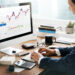 A person in a suit sits at a desk using a computer displaying financial charts and stock trading information, with a calculator, notebook, and coffee cup nearby. | 5StarsStocks