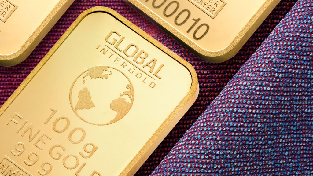 A close-up of gold bars labeled "GLOBAL INTERGOLD" and "100g FINE GOLD 999.9" resting on a textured purple and blue fabric. The bars feature a globe design and embossed serial numbers. | 5StarsStocks