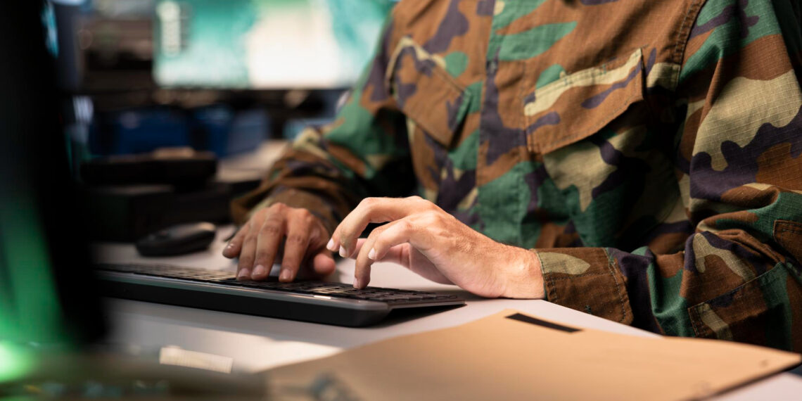 A person in a camouflage military uniform types on a computer keyboard at a desk, with a closed file folder in the foreground and multiple monitors in the background. | 5StarsStocks