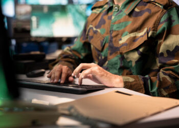 A person in a camouflage military uniform types on a computer keyboard at a desk, with a closed file folder in the foreground and multiple monitors in the background. | 5StarsStocks
