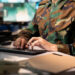 A person in a camouflage military uniform types on a computer keyboard at a desk, with a closed file folder in the foreground and multiple monitors in the background. | 5StarsStocks