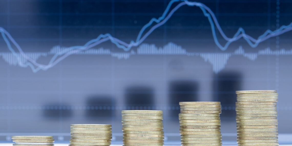 Stacks of coins arranged in ascending order in front of a blurry financial chart with rising and falling lines, symbolizing financial growth or investment trends. | 5StarsStocks