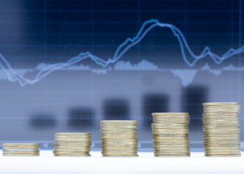 Stacks of coins arranged in ascending order in front of a blurry financial chart with rising and falling lines, symbolizing financial growth or investment trends. | 5StarsStocks