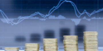 Stacks of coins arranged in ascending order in front of a blurry financial chart with rising and falling lines, symbolizing financial growth or investment trends. | 5StarsStocks