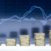 Stacks of coins arranged in ascending order in front of a blurry financial chart with rising and falling lines, symbolizing financial growth or investment trends. | 5StarsStocks