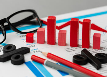 Red wooden bars, percentage symbols, pencils, and glasses are arranged on financial documents featuring charts and graphs. | 5StarsStocks