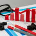 Red wooden bars, percentage symbols, pencils, and glasses are arranged on financial documents featuring charts and graphs. | 5StarsStocks