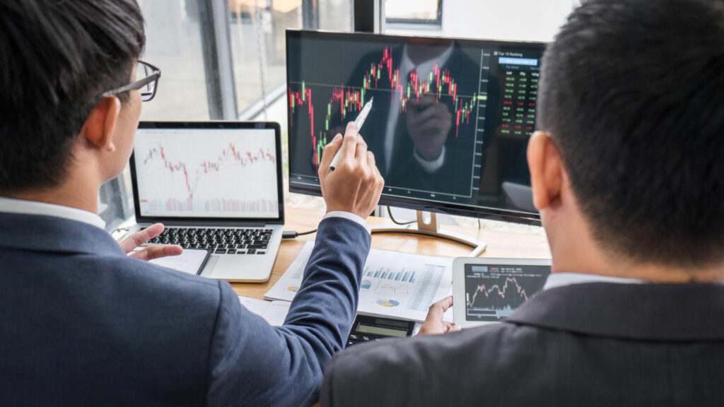 Two people in business attire analyze stock charts from 5StarsStocks.com on computer monitors and a laptop, discussing financial data and Stocks in an office setting with documents on the desk. | 5StarsStocks