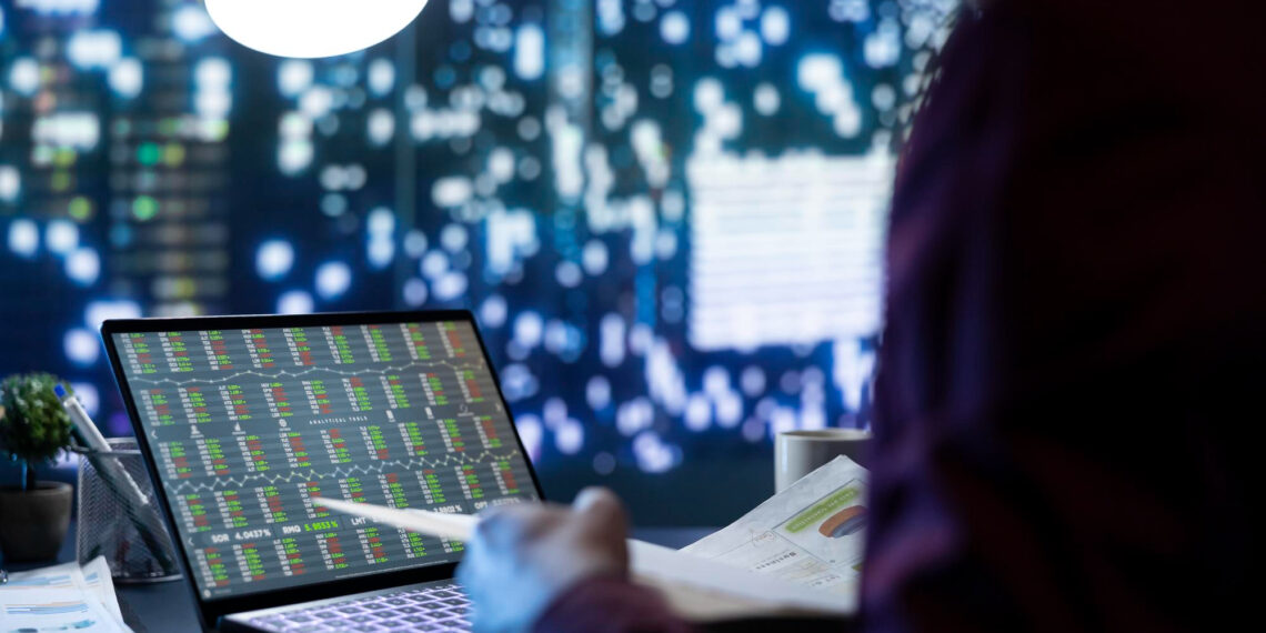 A person working at a desk views stock market data on a laptop screen, with city lights blurred in the background and a bright lamp overhead. | 5StarsStocks