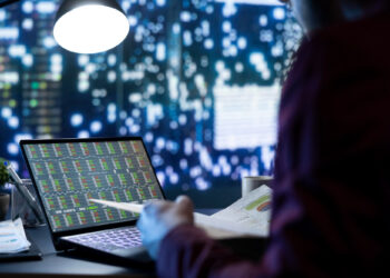A person working at a desk views stock market data on a laptop screen, with city lights blurred in the background and a bright lamp overhead. | 5StarsStocks