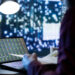 A person working at a desk views stock market data on a laptop screen, with city lights blurred in the background and a bright lamp overhead. | 5StarsStocks