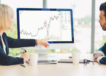 Two people in business attire sit at a desk, looking at a computer monitor displaying a rising stock chart. One person points at the screen while discussing the data. Coffee mugs and papers are on the table. | 5StarsStocks