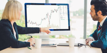 Two people in business attire sit at a desk, looking at a computer monitor displaying a rising stock chart. One person points at the screen while discussing the data. Coffee mugs and papers are on the table. | 5StarsStocks