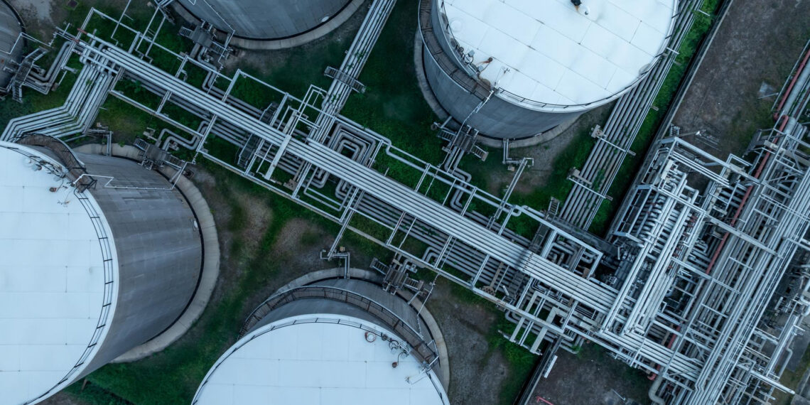 Aerial view of several large, round industrial storage tanks connected by a complex network of pipes on a grassy facility. | 5StarsStocks