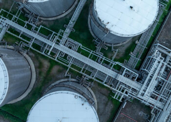 Aerial view of several large, round industrial storage tanks connected by a complex network of pipes on a grassy facility. | 5StarsStocks