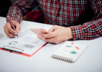 A person in a red plaid shirt uses a smartphone while writing on a document with charts. A notepad with colorful paperclips and a clipboard with papers are on the white desk. | 5StarsStocks
