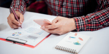 A person in a red plaid shirt uses a smartphone while writing on a document with charts. A notepad with colorful paperclips and a clipboard with papers are on the white desk. | 5StarsStocks