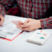 A person in a red plaid shirt uses a smartphone while writing on a document with charts. A notepad with colorful paperclips and a clipboard with papers are on the white desk. | 5StarsStocks