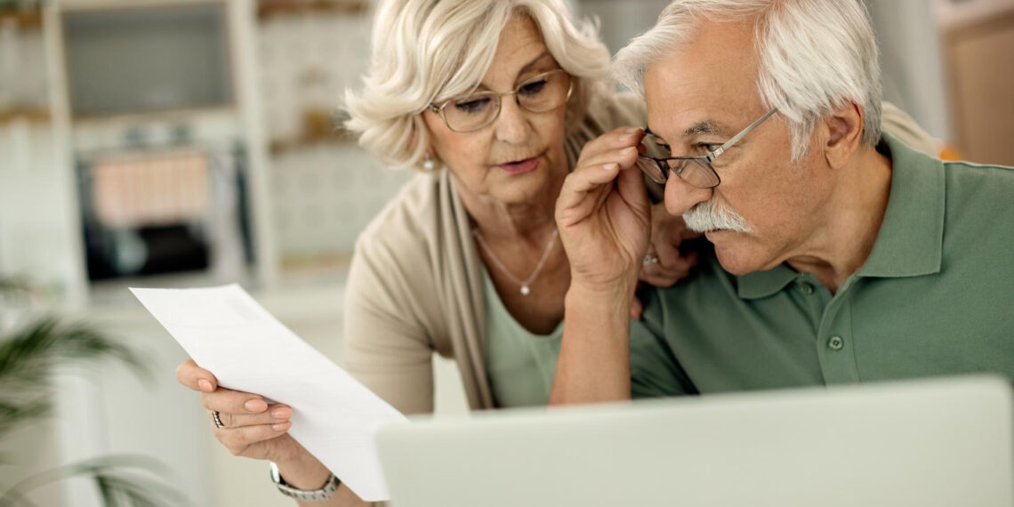 An elderly couple looks at a laptop together while the woman holds a sheet of paper, both appearing focused and concerned in a bright home setting. | 5StarsStocks