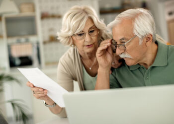 An elderly couple looks at a laptop together while the woman holds a sheet of paper, both appearing focused and concerned in a bright home setting. | 5StarsStocks