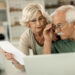 An elderly couple looks at a laptop together while the woman holds a sheet of paper, both appearing focused and concerned in a bright home setting. | 5StarsStocks