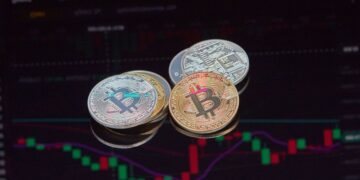Three physical Bitcoin tokens in gold, silver, and copper are placed on a surface with a blurred cryptocurrency trading chart with green and red candlesticks displayed in the background. | 5StarsStocks
