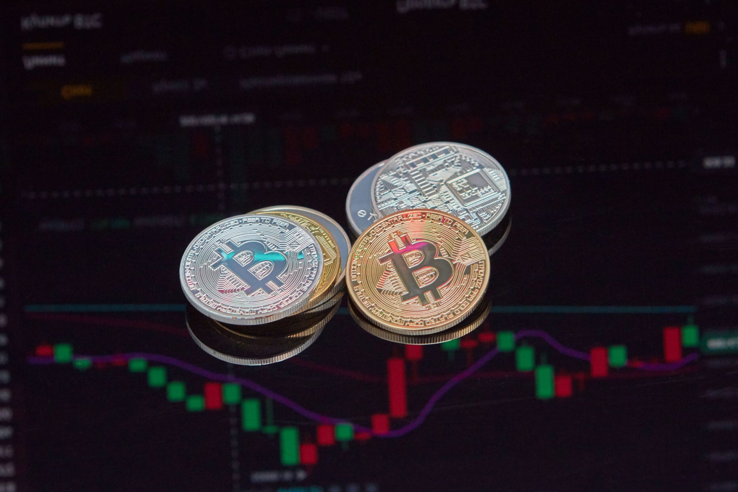 Three physical Bitcoin tokens in gold, silver, and copper are placed on a surface with a blurred cryptocurrency trading chart with green and red candlesticks displayed in the background. | 5StarsStocks