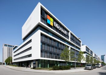 A modern Microsoft office building with a large logo on the facade, surrounded by trees and parked cars under a clear blue sky. | 5StarsStocks