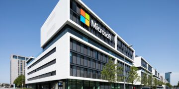 A modern Microsoft office building with a large logo on the facade, surrounded by trees and parked cars under a clear blue sky. | 5StarsStocks