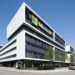 A modern Microsoft office building with a large logo on the facade, surrounded by trees and parked cars under a clear blue sky. | 5StarsStocks